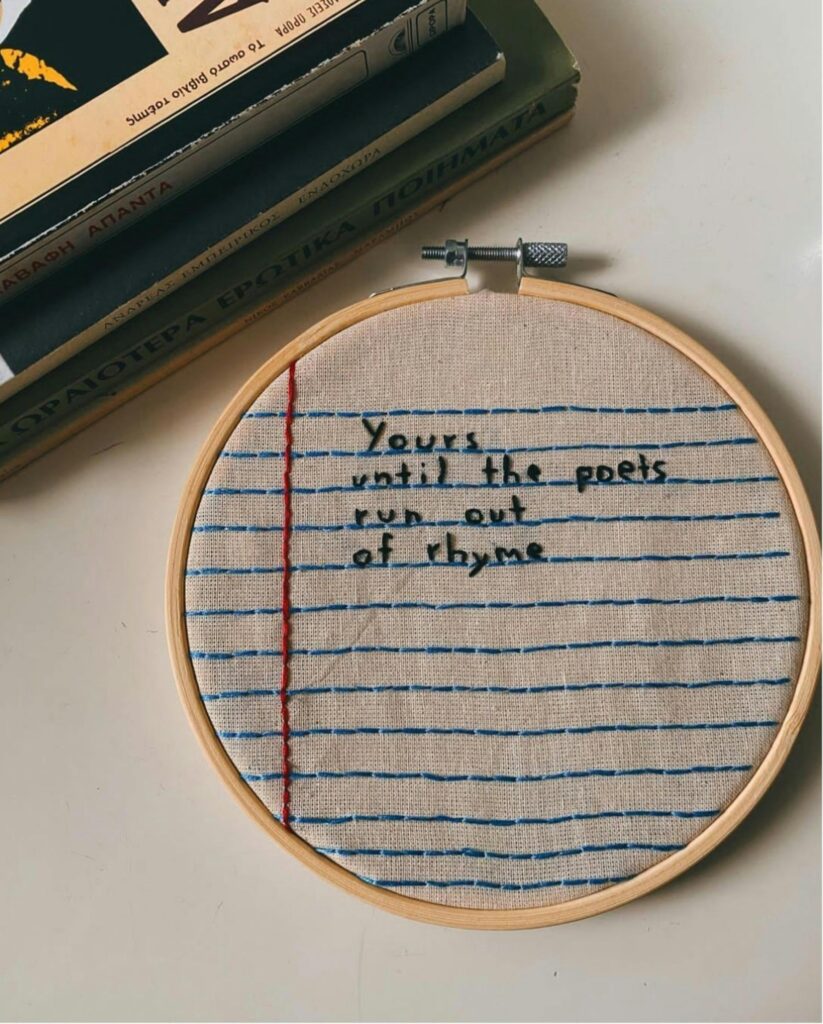 Embroidery hoop with text on notebook-style fabric, beside stacked books.